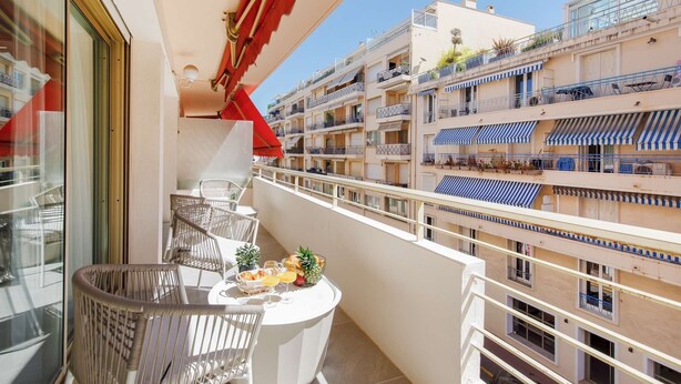 En balkon med fletstole og et bord dækket med frugt og drikkevarer, med udsigt over en bygning med flere etager, balkoner og stribede markiser.
