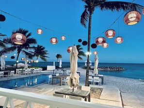 Zona junto a la piscina con mesas y sillas, palmeras y luces colgantes.