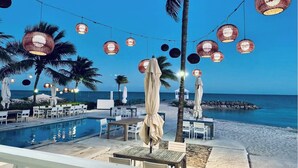 Zona junto a la piscina con mesas y sillas, palmeras y luces colgantes.