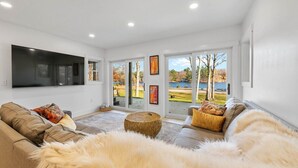Ein Wohnzimmer mit großem Flachbildfernseher, einer Sofagarnitur, einem flauschigen Teppich und Blick auf einen See und Bäume durch große Fenster.