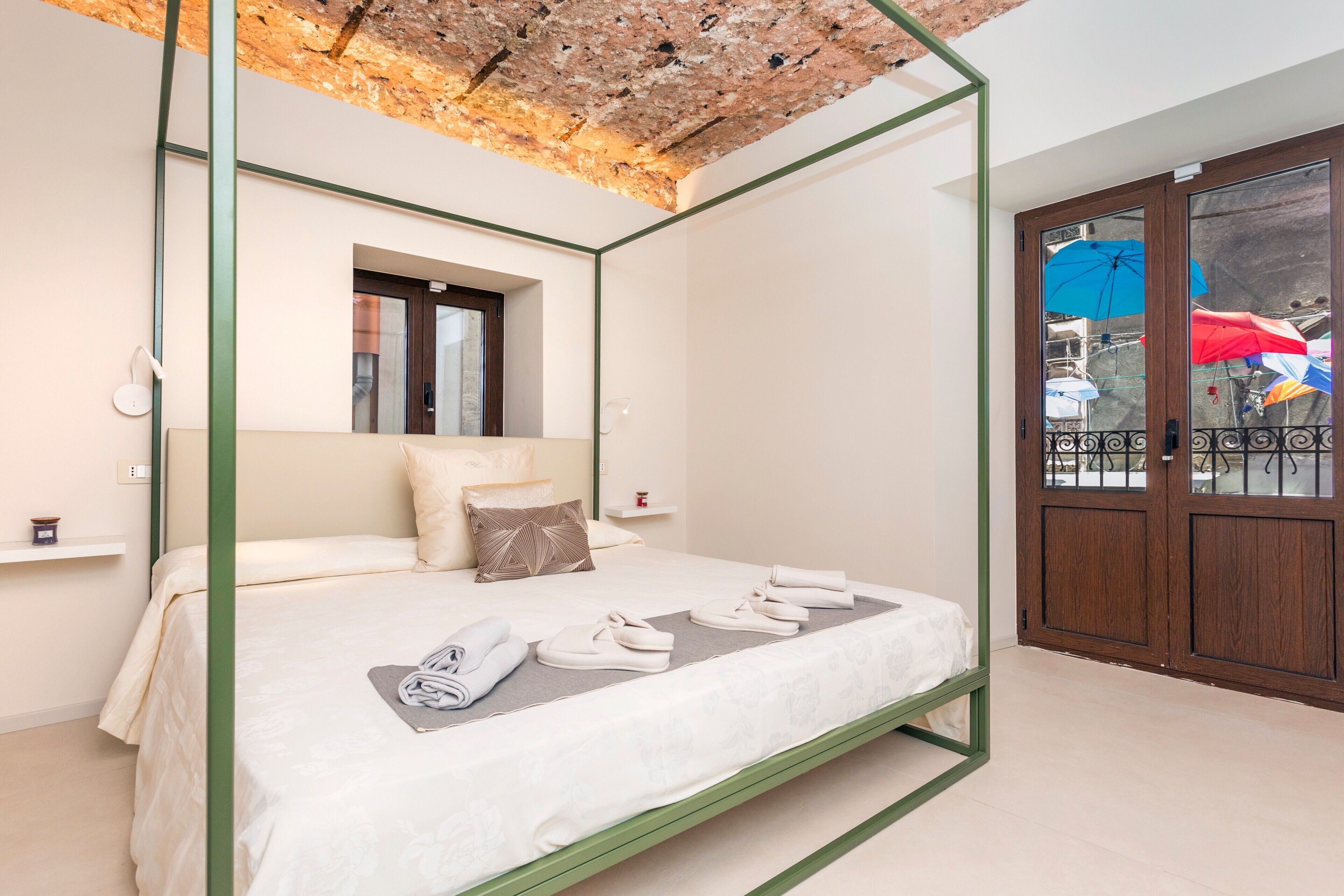 Una cama con dosel de estructura verde, una ventana encima y vista a un balcón con sombrillas.