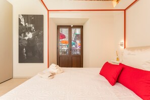 Una camera da letto con un letto, un cuscino rosso, un cuscino bianco, un'opera d'arte a parete, una porta con un pannello di vetro e un comodino con una bottiglia.
