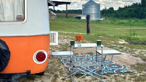 A vintage camper parked on a grassy field with a picnic setup including a table, chairs, and a patterned tablecloth.