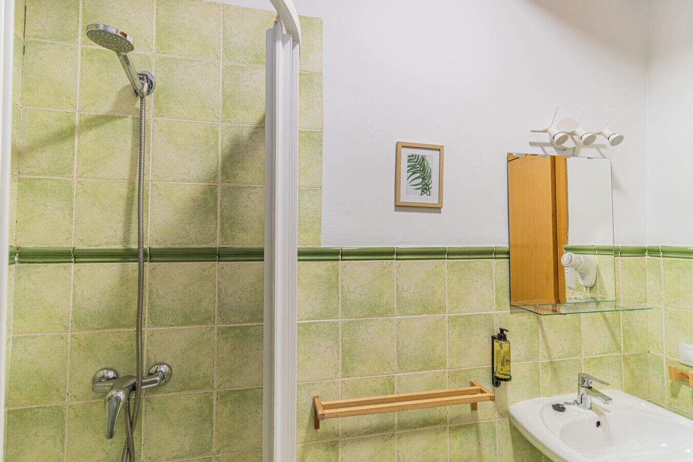 Baño con azulejos verdes, ducha, lavamanos, toallero de madera y planta en marco.