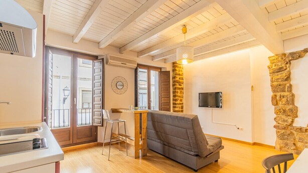 Un vaste espace de vie agrémenté d’un mur en pierre, d’un téléviseur à écran plat fixé au mur, d’un plafond en bois et d’un coin cuisine équipé d’un évier et d’une cuisinière.