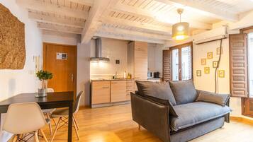 A modern living room with a sofa, a dining table with chairs, a kitchen area, and a large window with shutters.
