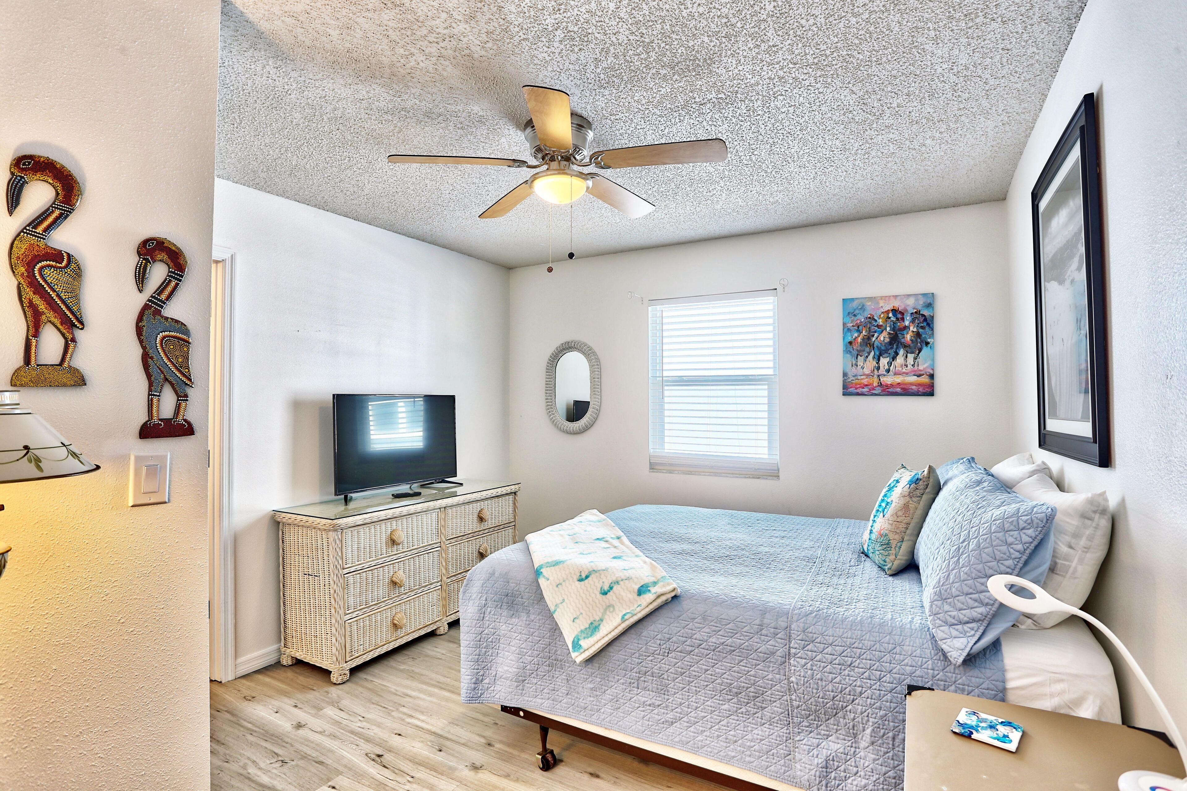 A bedroom with a bed, dresser, TV, ceiling fan, artwork, and a window.