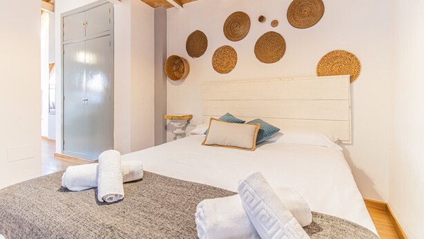 Une chambre avec un plafond en bois, un lit avec une literie blanche, une tête de lit ornée de motifs tissés et un tapis gris.
