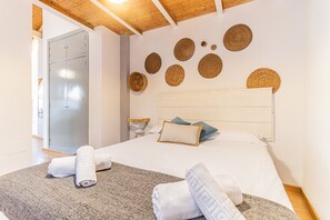 Un dormitorio con techo de madera, cama con ropa blanca, cabecera con adornos tejidos y alfombra gris.