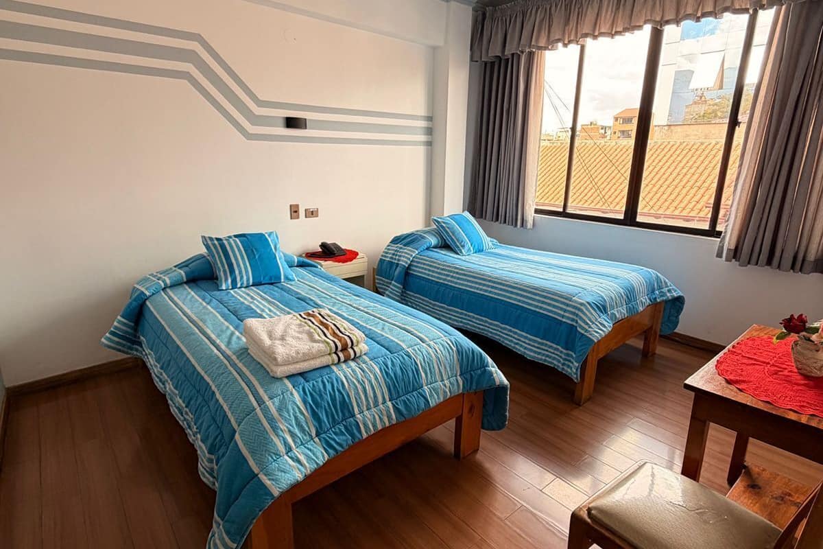 Une chambre d’hôtel avec deux lits, un plancher en bois, une petite table recouverte d’une nappe rouge et une fenêtre avec des rideaux.