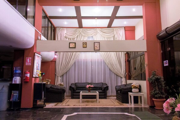 A hotel lobby with a black leather sofa set, a small coffee table, and a decorative vase with flowers. There are two framed certificates on the wall, a fire extinguisher, and a water dispenser.
