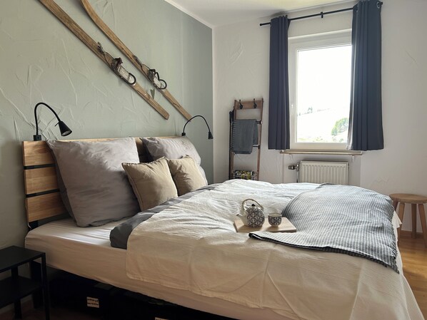 Cama bem arrumada com cabeceira, mesa de cabeceira, janela com cortinas e radiador.