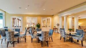 A spacious dining area with round tables and chairs, large windows, and a warm color scheme. - Natchez Grand Hotel (Natchez)