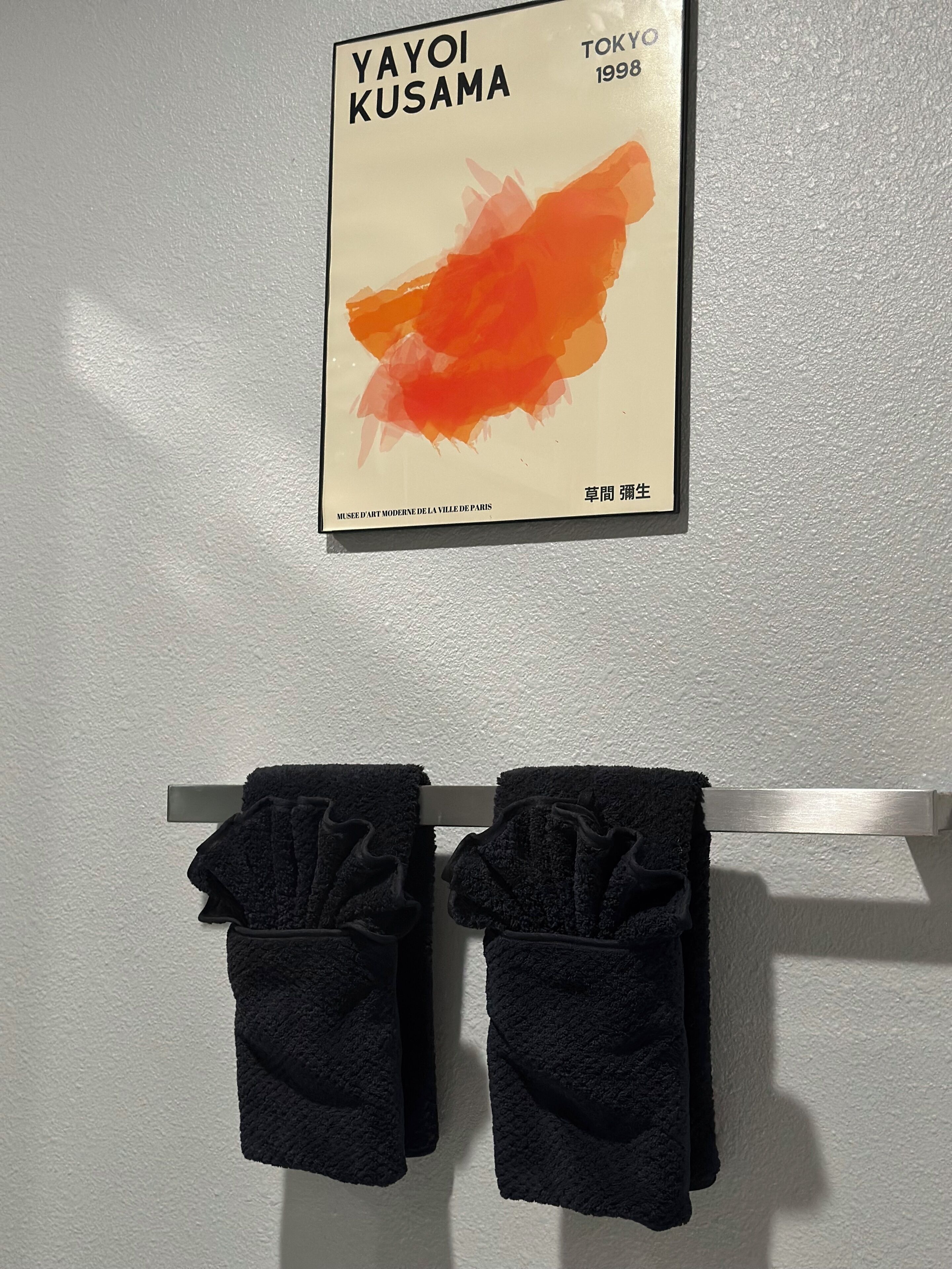 Kehyksiin asetettu taideteos, jonka nimi on ”YAYOI KUSAMA” ja joka on vuodelta 1998, on esillä valkoisella, kuvioidulla seinällä. Sen alla on metallitelineessä kaksi tummaa pyyhettä.