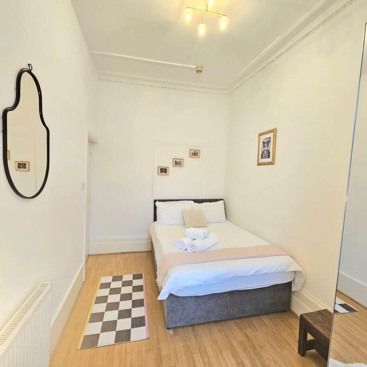 A small, clean room with a bed, a checkered rug, a mirror, and a ceiling light fixture.