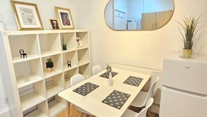 Un espace de vie compact comprenant une table à manger, des chaises, un réfrigérateur blanc, un mur miroir et une étagère blanche agrémentée d’objets décoratifs.