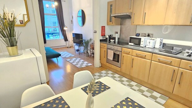 Une cuisine moderne avec des meubles en bois, un four à micro-ondes et une cuisinière. On y trouve un coin repas avec une table et des chaises, et une vue sur la ville grâce à de grandes fenêtres.