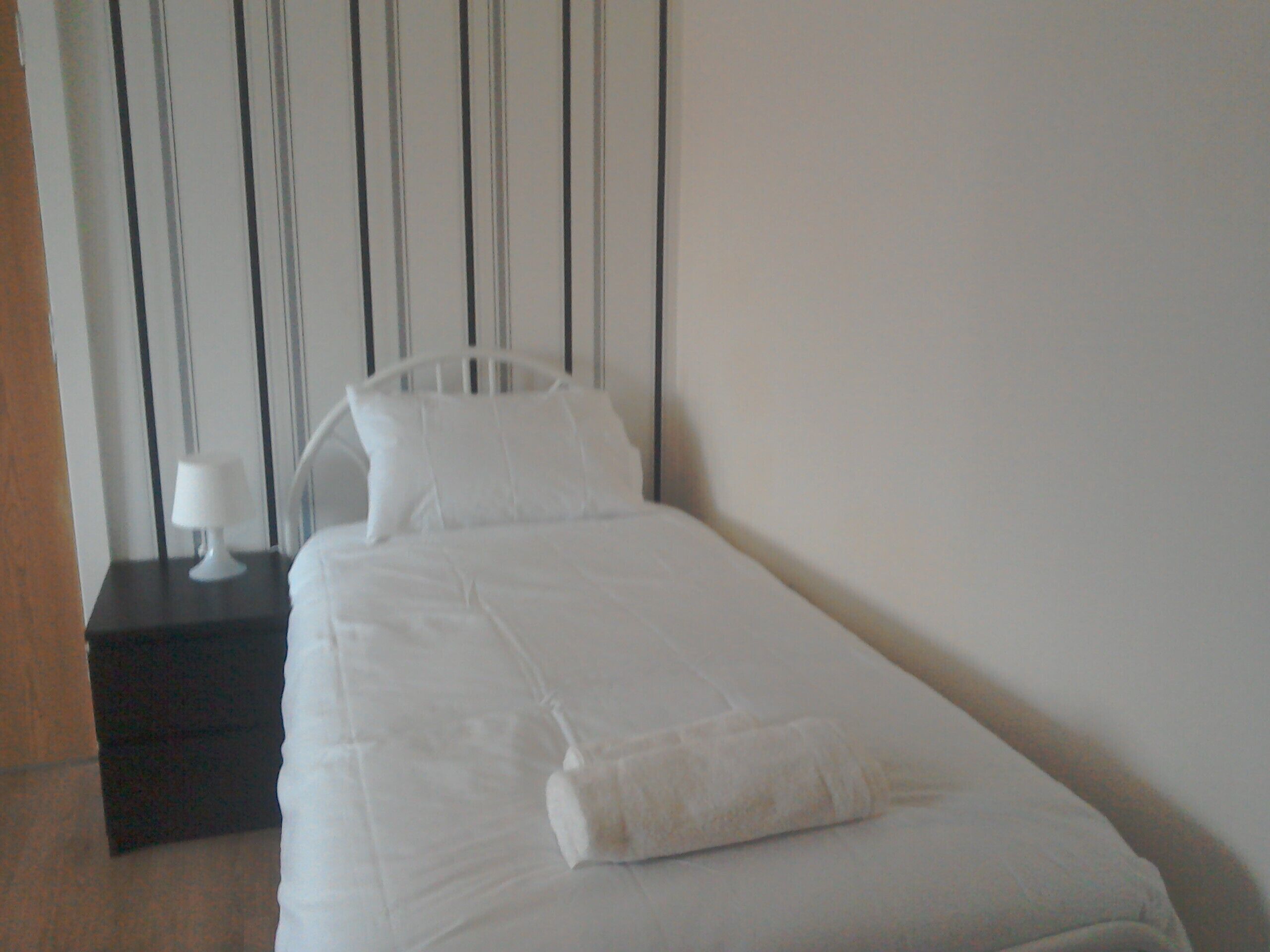 Un petit lit bien fait, avec du linge de lit blanc et des oreillers, une table de chevet avec une lampe, et un mur à rayures en arrière-plan.