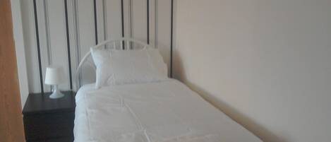 Un petit lit bien fait, avec du linge de lit blanc et des oreillers, une table de chevet avec une lampe, et un mur à rayures en arrière-plan.