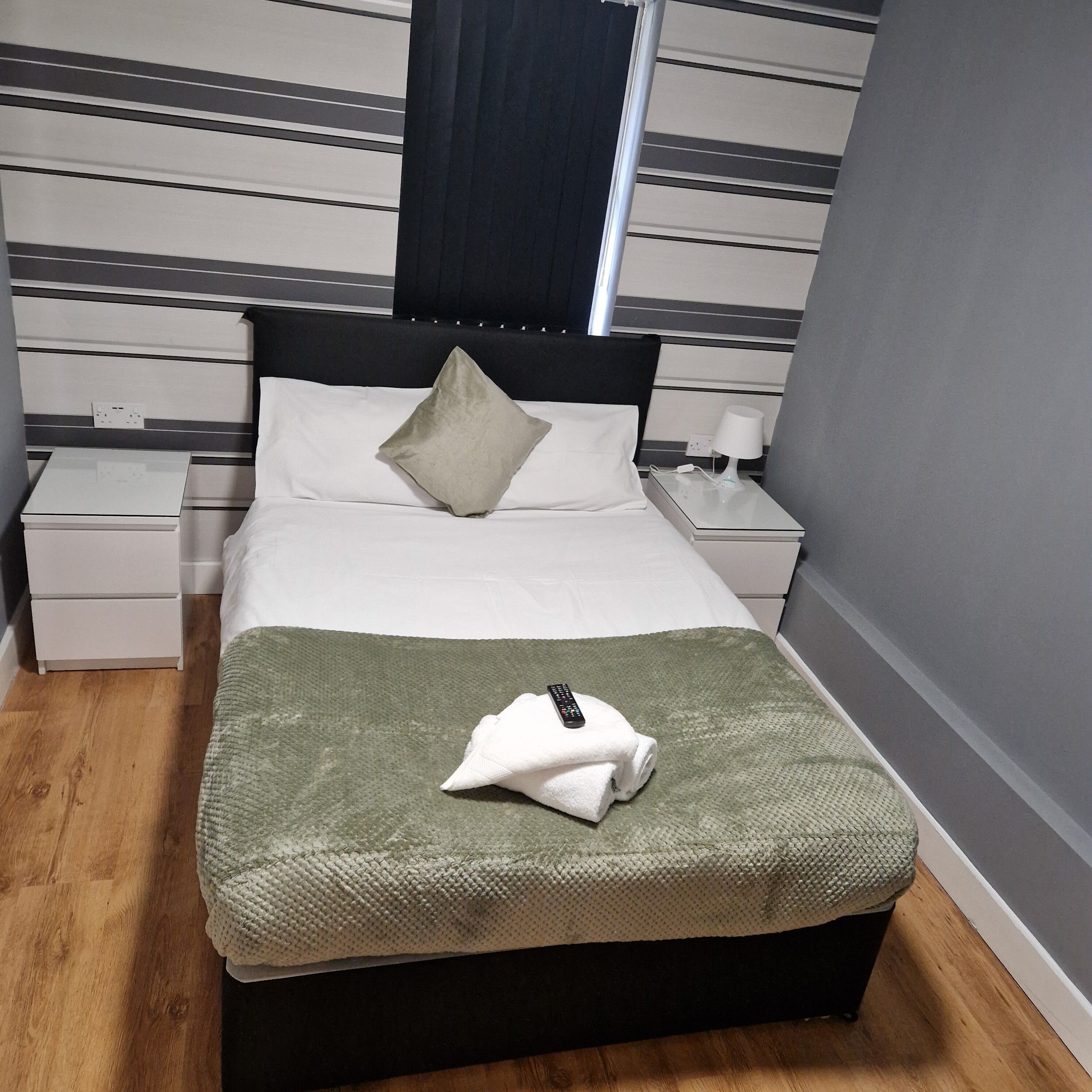 A neatly made bed with a green blanket, a pillow, and a remote control on it. There is a nightstand with a lamp and a power outlet. The room has a striped wall and a window with curtains.