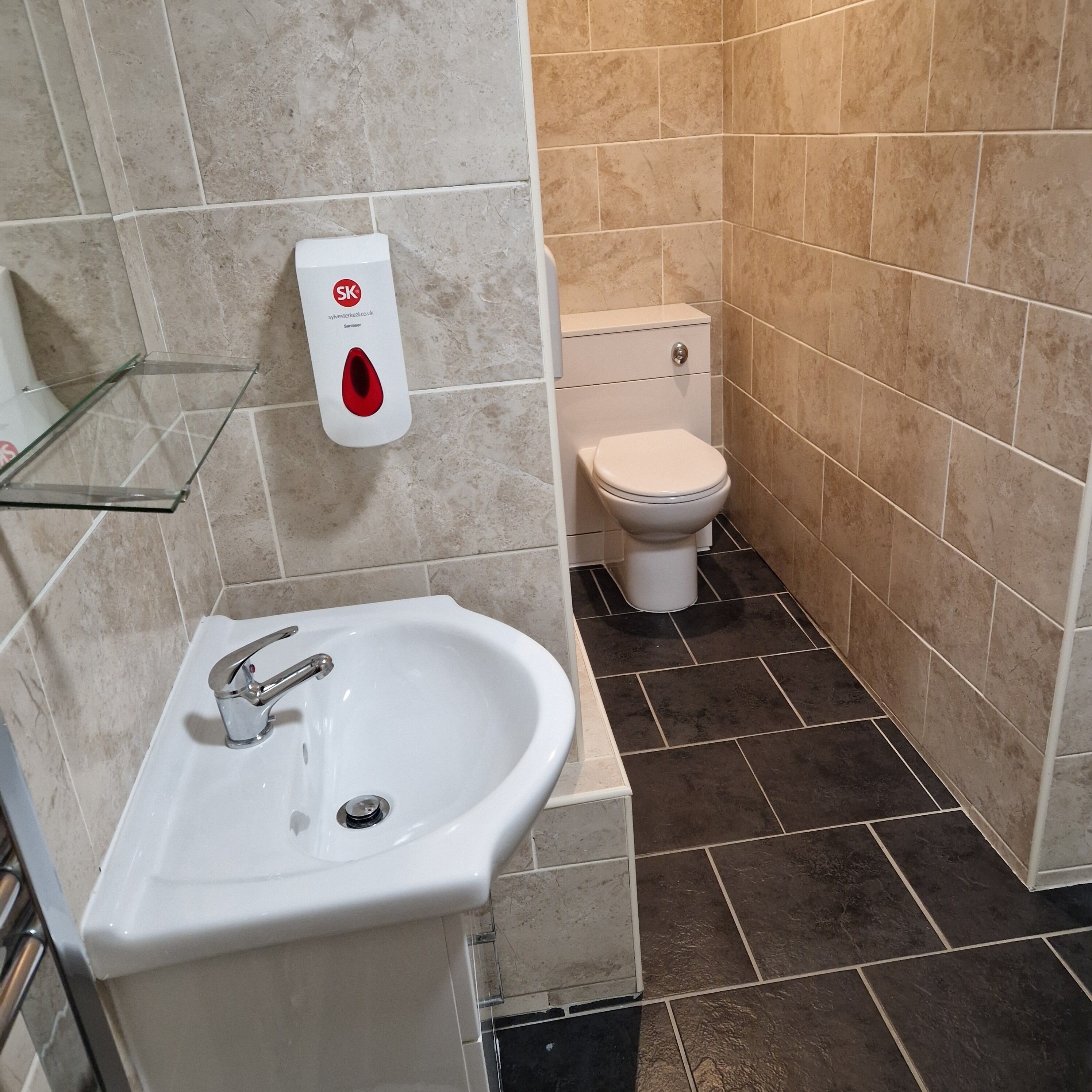 Een badkamer met een wit wastafel, een glazen plankje, een toilet en een handdesinfecteerder.