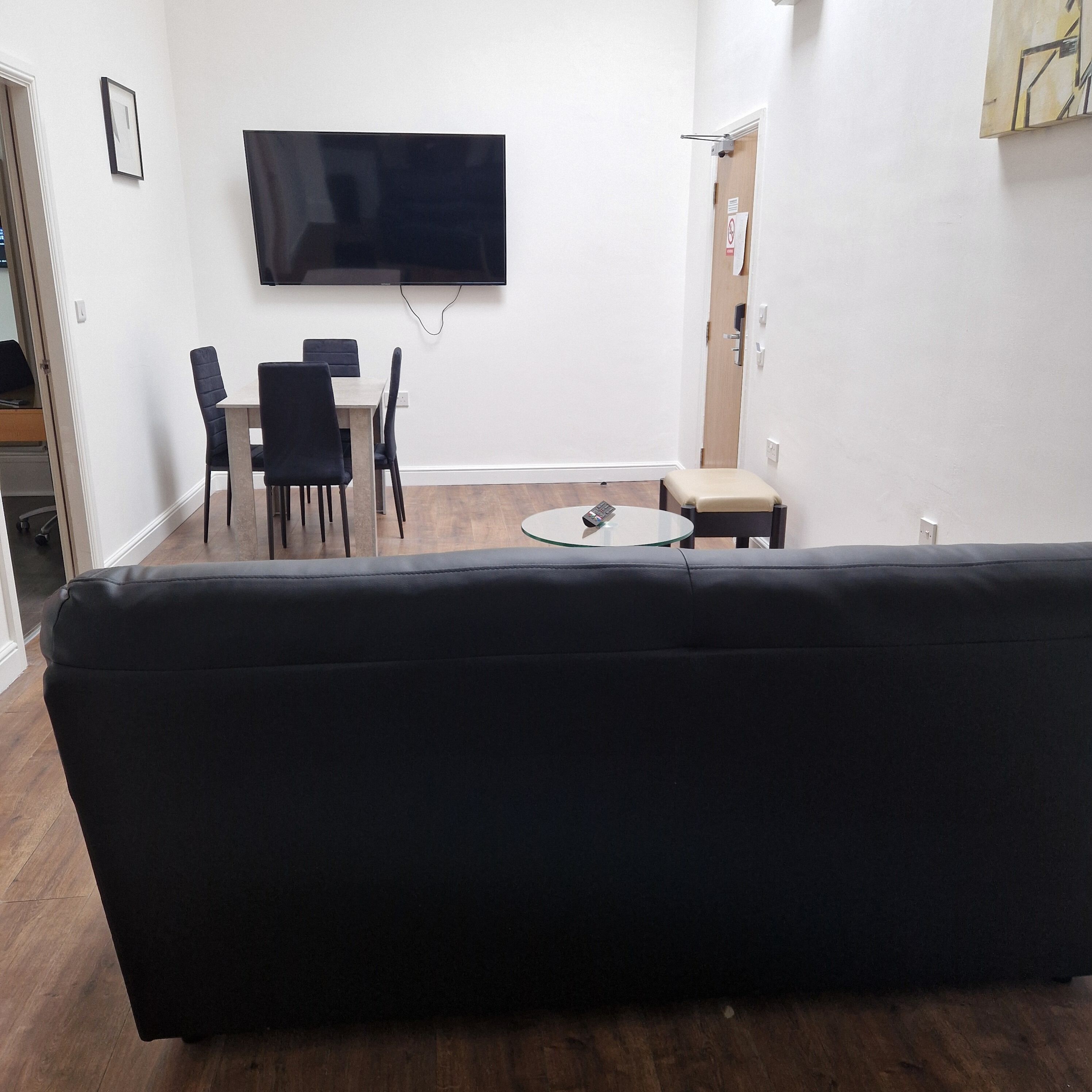 A living room with a black sofa, a TV mounted on the wall, a dining table with chairs, and a small round table with a remote control.