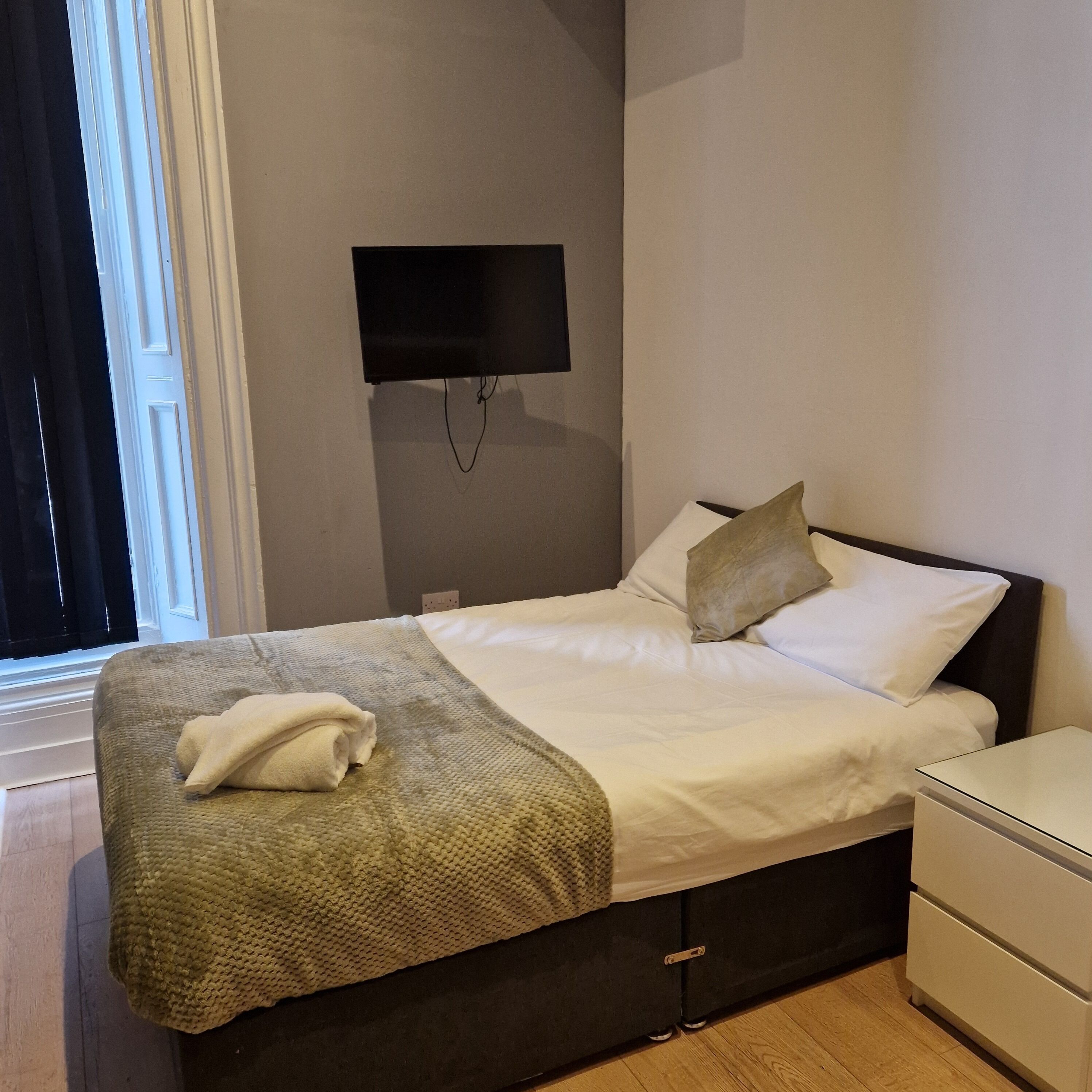 Una cama bien tendida con edredón blanco y almohadas, una toalla doblada sobre la cama, cabecera oscura, mesita de noche y un televisor de pantalla plana montado en la pared.