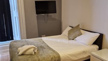 Una cama bien tendida con edredón blanco y almohadas, una toalla doblada sobre la cama, cabecera oscura, mesita de noche y un televisor de pantalla plana montado en la pared.