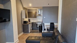 A modern living room with a sectional sofa, a flat-screen TV mounted on the wall, and a kitchen in the background. - The Dorchester Hotel (Hull)