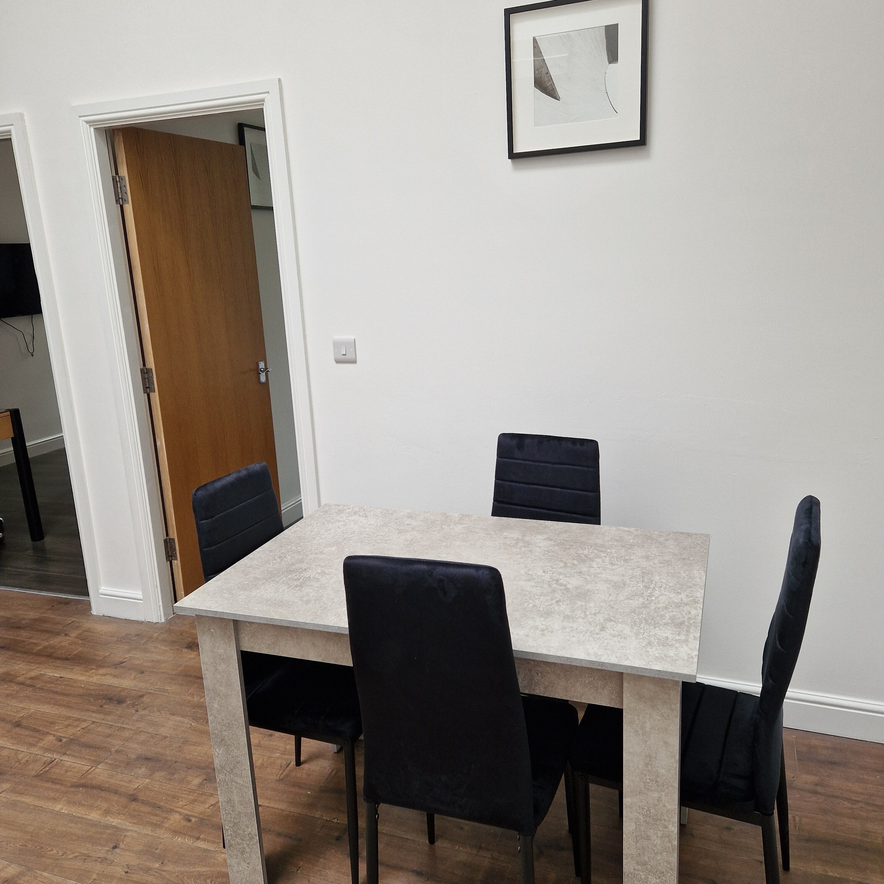 A dining area with a table and four chairs, a framed picture on the wall, and a wooden door leading to another room.