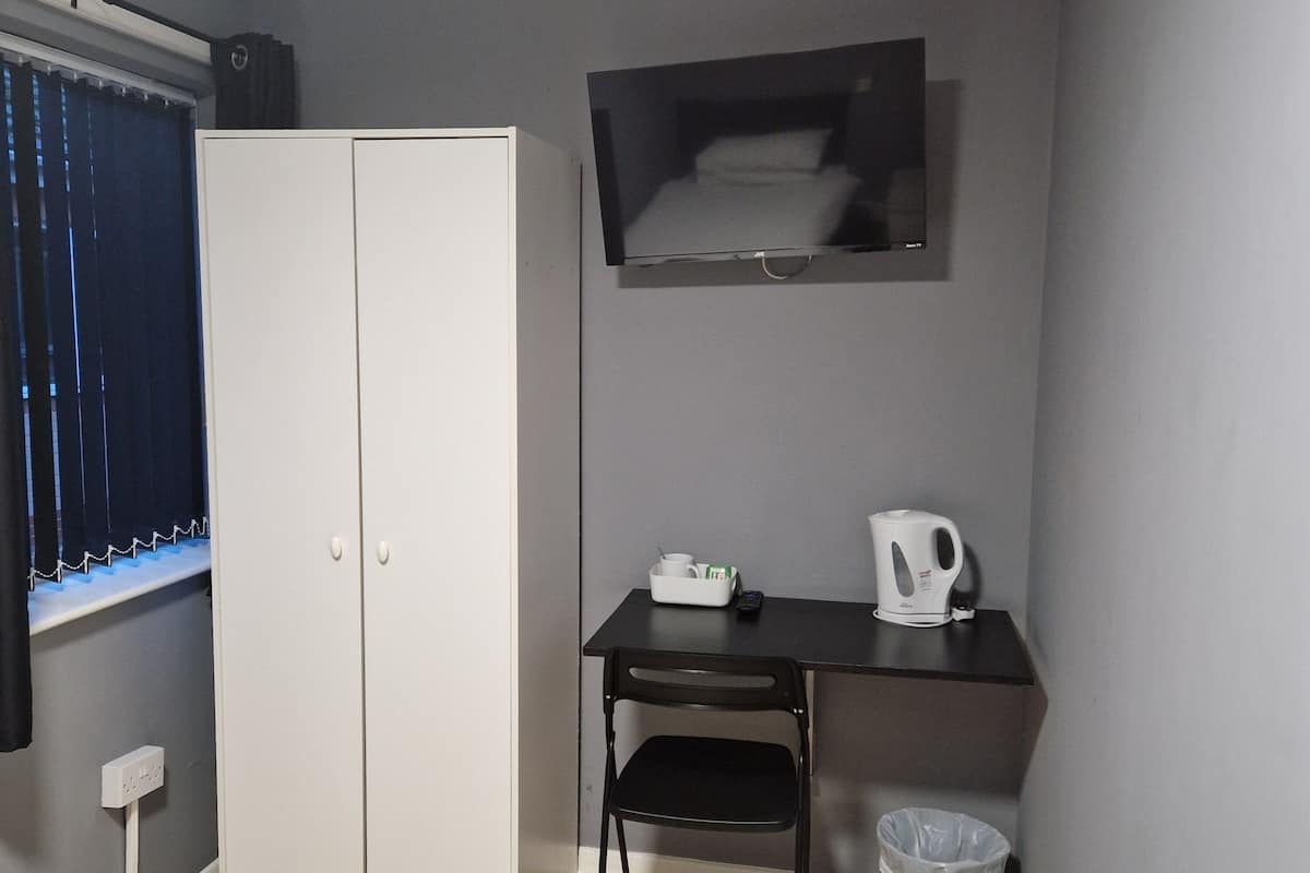 A room with a wardrobe, a desk with a kettle, a chair, and a trash bin.