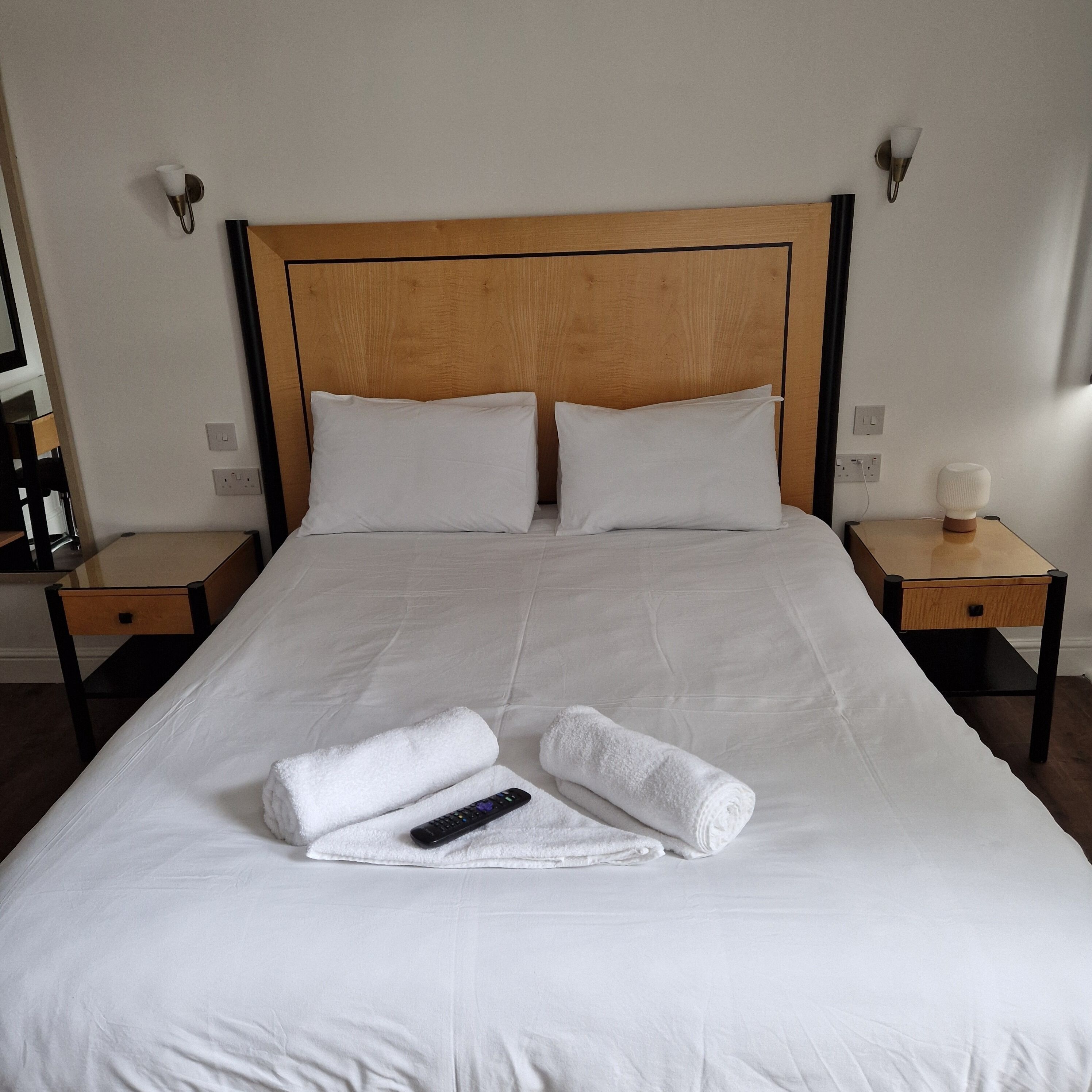 A neatly made bed with white linens, two pillows, and two rolled towels. There is a remote control on one of the towels. The room has wooden headboard and nightstands with lamps.