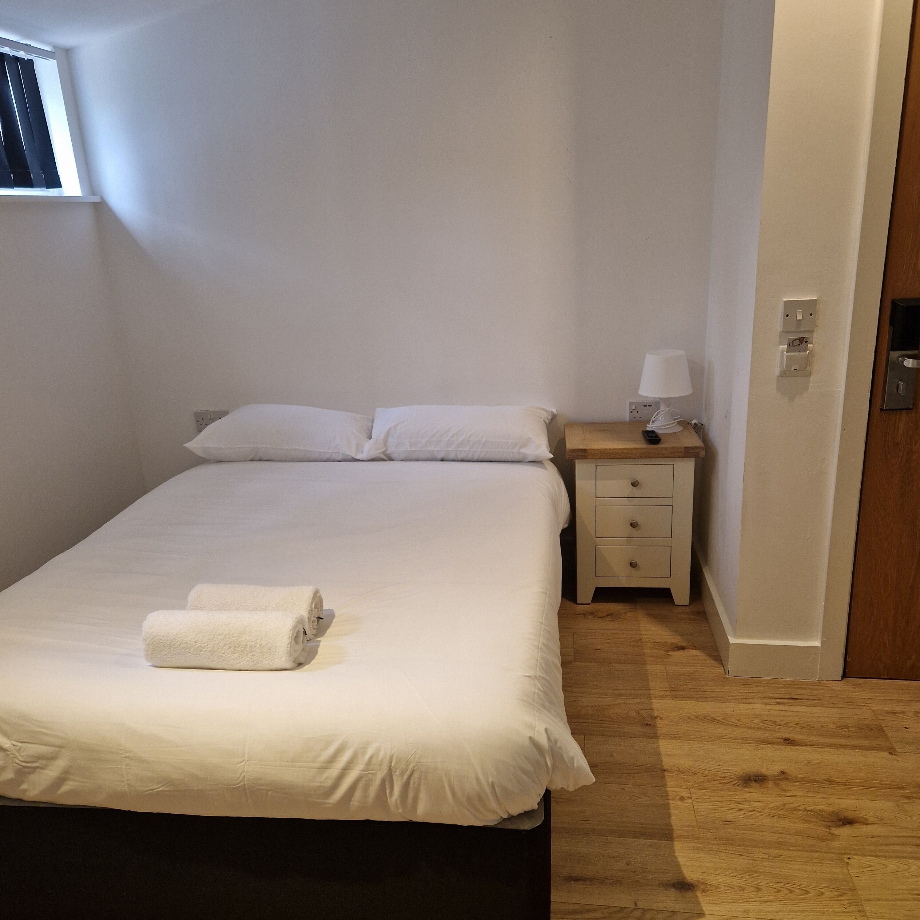 A single bed with white linens, a wooden nightstand with a lamp, and a power outlet.
