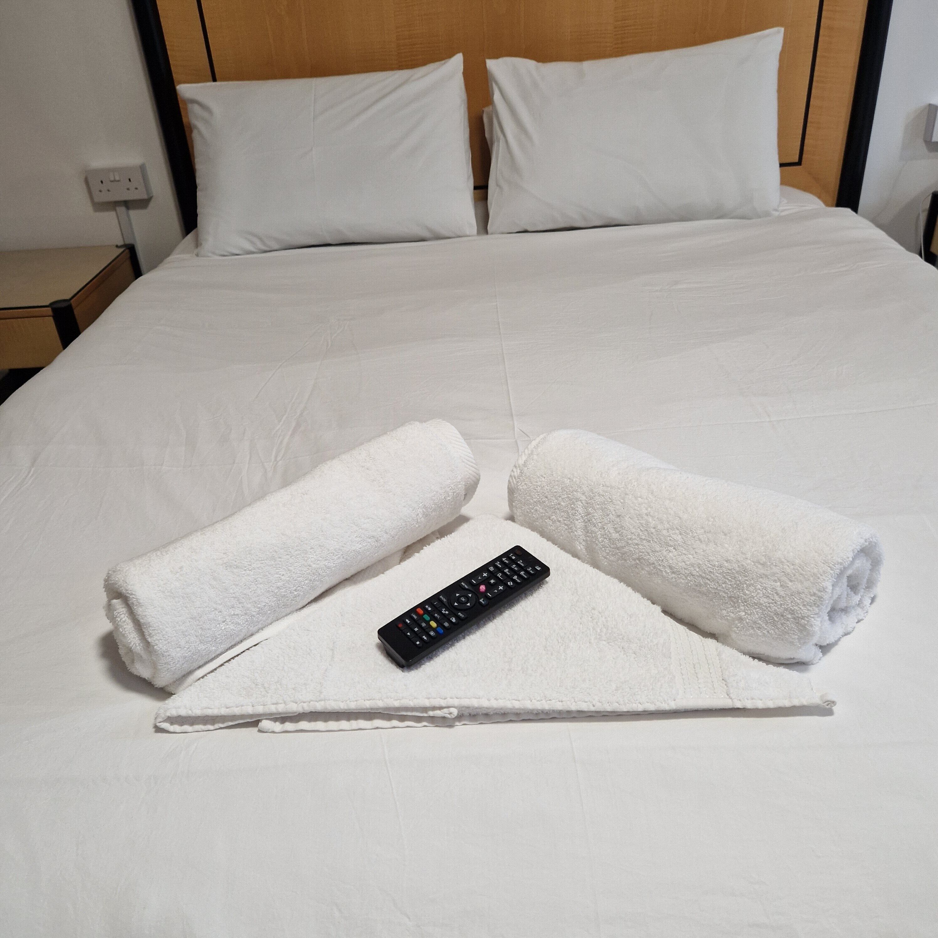 A neatly made bed with white linens, two rolled towels, and a remote control on the bed.