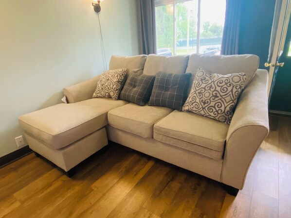 A beige sectional sofa with patterned cushions, a wall-mounted light, and a view of the outdoors through a window. - WI river, Kash Park Play & Stay, Pet friendly (Stevens Point)