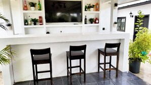 Een barruimte met drie houten barkrukken, een flatscreen televisie erboven en planken met flessen en planten.