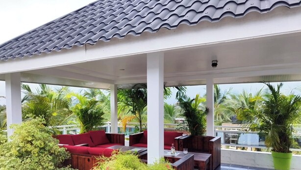 Een overdekt terras met rode banken en houten tafels, omgeven door weelderige begroeiing en palmbomen.