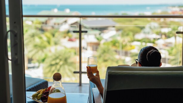 Une personne est assise près d’une fenêtre, avec vue sur une zone côtière. Elle tient un verre à la main, et sur la table, on peut voir une assiette de fruits et une bouteille.