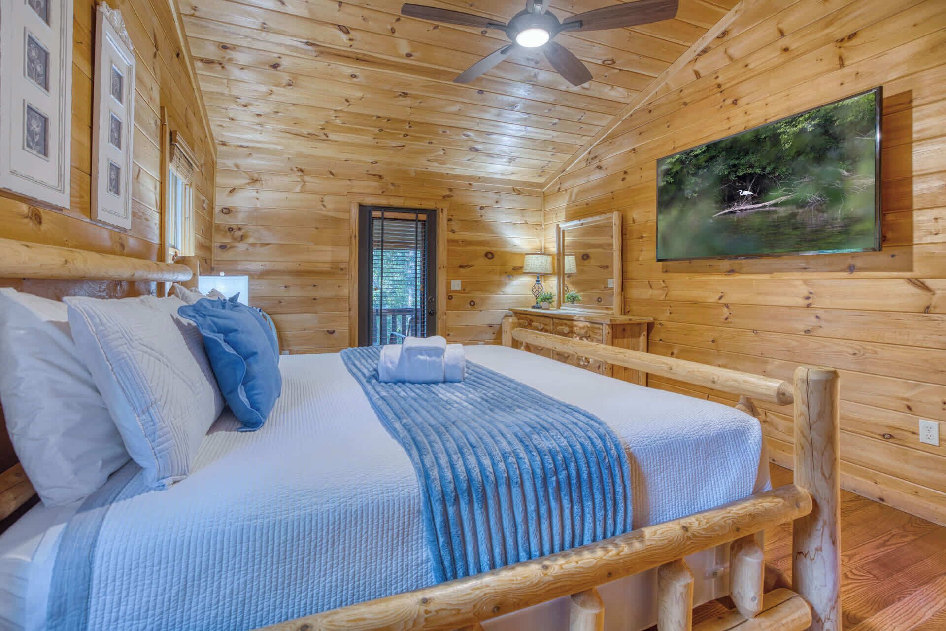 A wooden cabin bedroom with a bed, a ceiling fan, a window, and a painting on the wall.