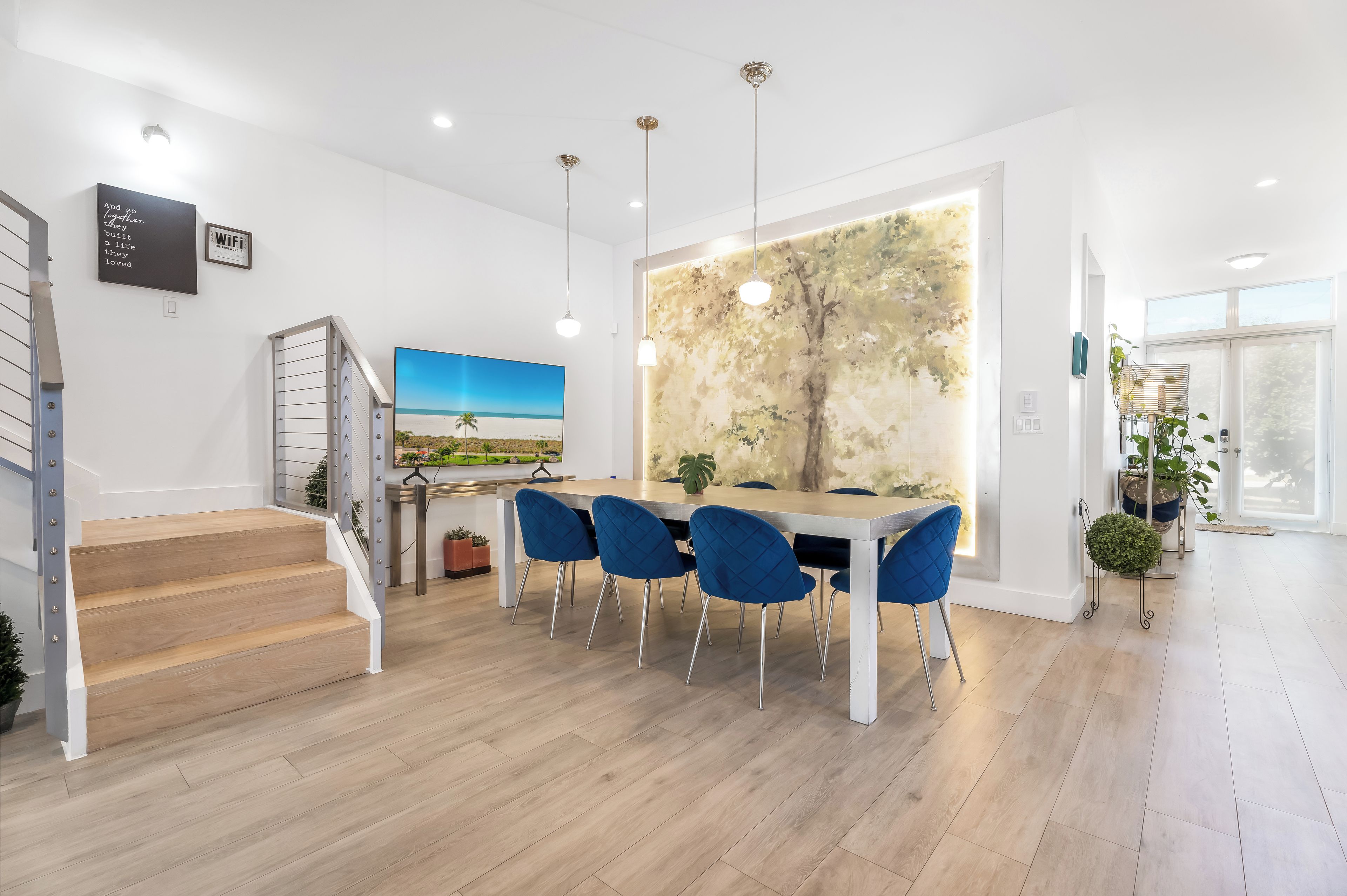 A modern dining area with a wooden staircase, a dining table with blue chairs, and a large wall mural.