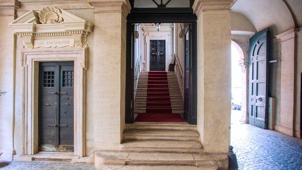 A grand staircase with a red carpet leading to a dark wooden door, flanked by ornate columns and a decorative ceiling. - Palazzo Costaguti – La Dimora del Tempo (Roma)