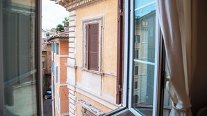 A narrow street view through a window with a building featuring a balcony with potted plants and a door. - Palazzo Costaguti – La Dimora del Tempo (Roma)
