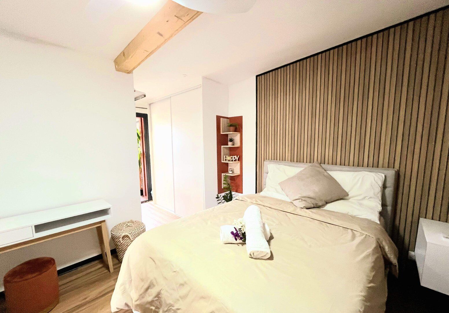 Une chambre à coucher avec un lit, une tête de lit en bois, une petite étagère avec des plantes et un mur blanc.