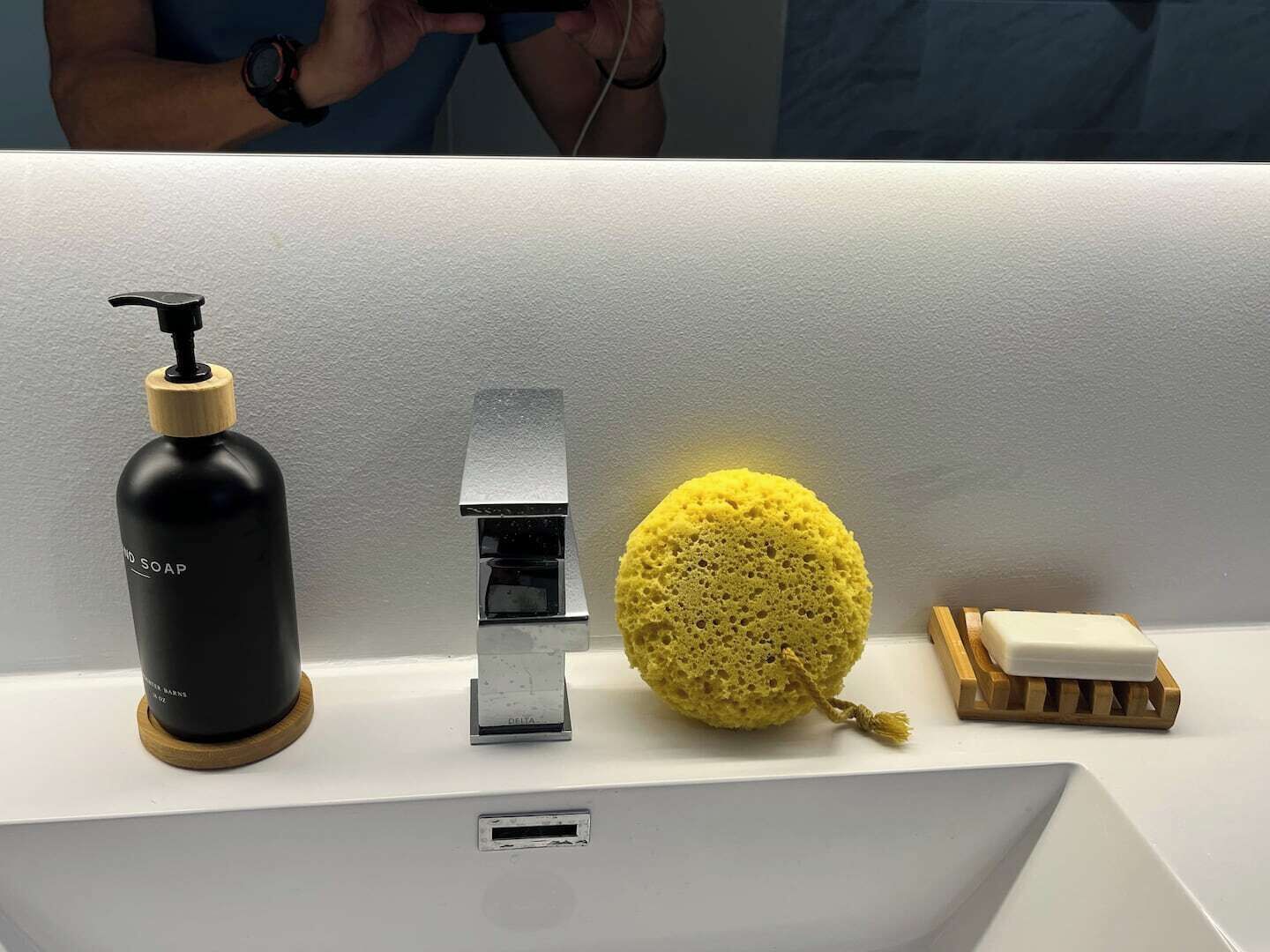 A bathroom sink with a black soap dispenser, a silver faucet, a yellow sponge, and a soap bar on a wooden holder.