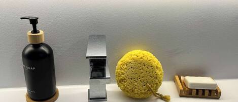 A bathroom sink with a black soap dispenser, a silver faucet, a yellow sponge, and a soap bar on a wooden holder.