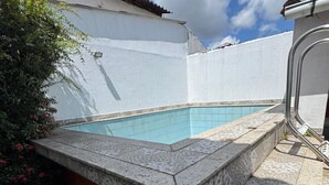 A swimming pool with a tiled edge, surrounded by a brick wall and a white wall with a tiled roof.