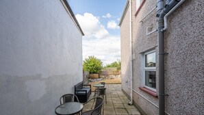 A narrow outdoor patio with a seating area consisting of a bench and several chairs, a small round table, and a window on the right side. - Emerald House by Tŷ SA (Newport)