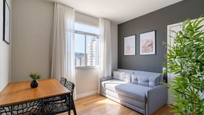 Una sala de estar moderna con un sofá gris, una mesa de comedor de madera con sillas, una planta en maceta y vistas a la ciudad a través de un ventanal grande.