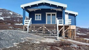 Una cabaña azul con terraza de madera y escaleras, rodeada por un paisaje montañoso.