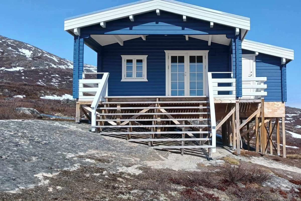 A blue cabin with a wooden deck and stairs, surrounded by a mountainous landscape.